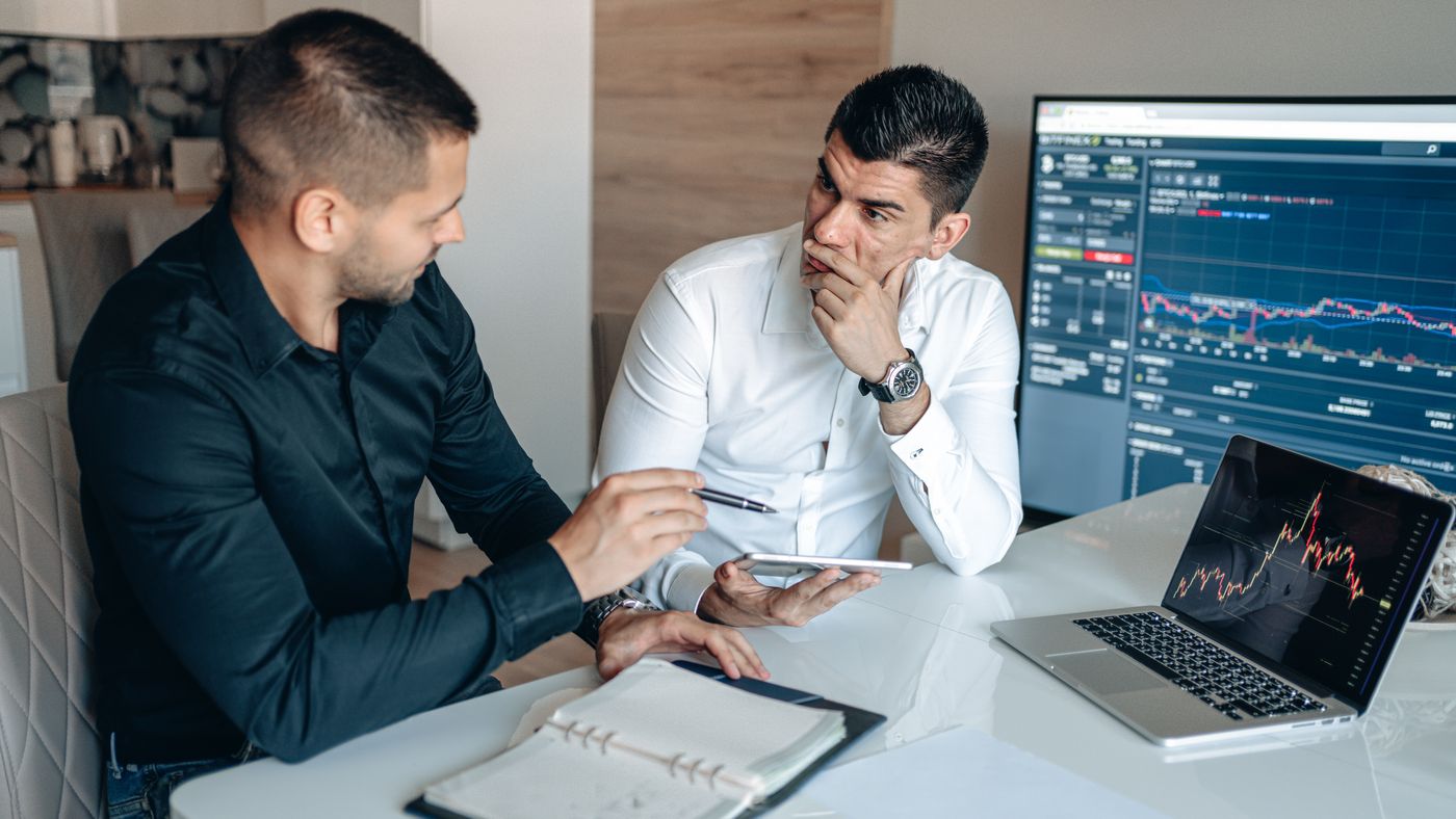 Two traders discussing market charts on a trading monitor and laptop